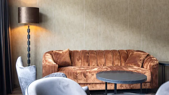 En orange sammetssoffa står tom i ett mjukt belyst rum, tillsammans med en golvlampa och omgiven av grå stolar och runda bord mot en beige vägg.