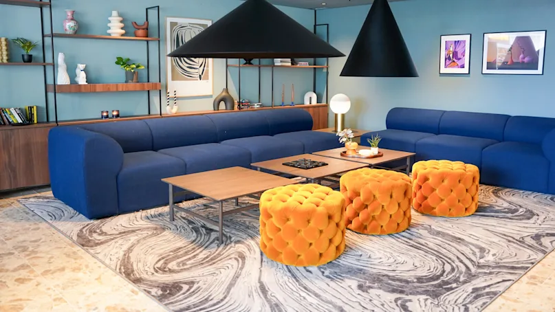 Blue sofas sit on a swirling-patterned rug, while orange, tufted stools are positioned around wooden tables; black cone-shaped lights hang above in a modern lounge space.