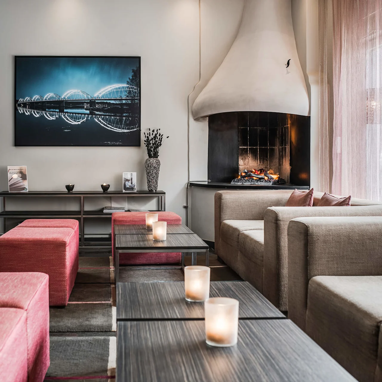 En modern lounge med beige soffor och röda pallar runt små bord med ljus. En öppen spis ger värme. En inramad bild av en bro och broschyrer dekorerar väggarna. 