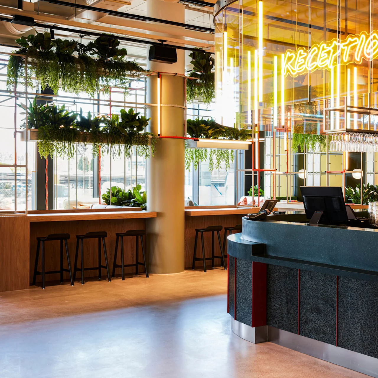 Reception desk med planter og neonskilt, der siger "RECEPTION"; levende moderne indstilling med hængende glas og skamler nær vinduer med naturligt lys.