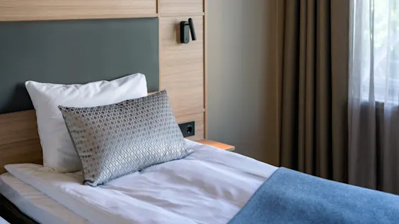 A neatly made bed with white linens and a blue blanket in a modern room. A decorative pillow rests on top. Wall-mounted lights illuminate a wooden headboard next to a curtain-covered window.