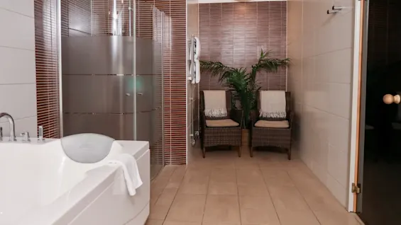 A white bathtub with a towel draped over the edge stands beside a frosted glass shower. Two wicker chairs with cushions are placed against brown tiled walls.