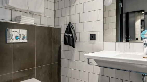 Toalett monterad på kaklad vägg med två vikta handdukar ovanför; elegant, vit handfat till höger med spegel som reflekterar dörröppning; grå och vita kakel skapar en modern badrumsmiljö.