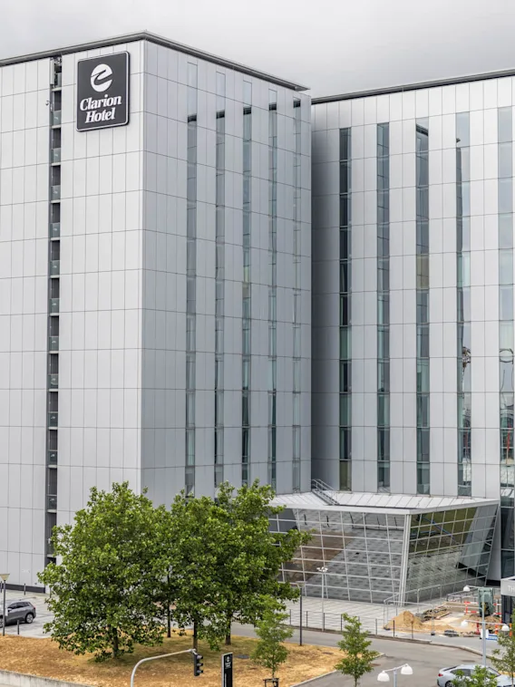 En høj, moderne bygning mærket “Clarion Hotel” med store glasvinduer står i et urbant miljø. Flagene vejrer nær indgangen.
