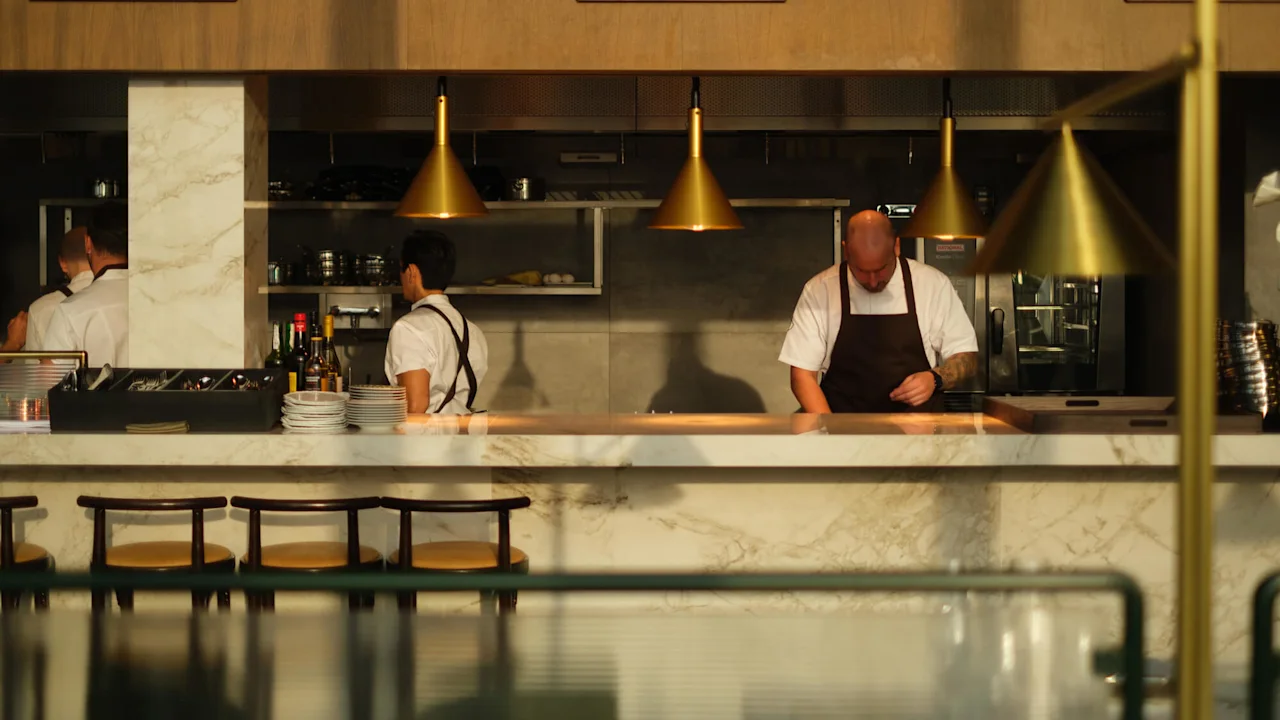 En kokk tilbereder mat i et moderne kjøkken med marmordisker og overlys. Andre kokker jobber i bakgrunnen, omgitt av hyller og ingredienser.