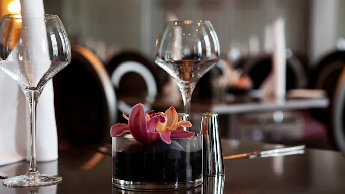 A wine glass stands on a restaurant table, reflecting soft light. Beside it, an orchid floats in a small glass pot. Other tables and chairs fill the background.