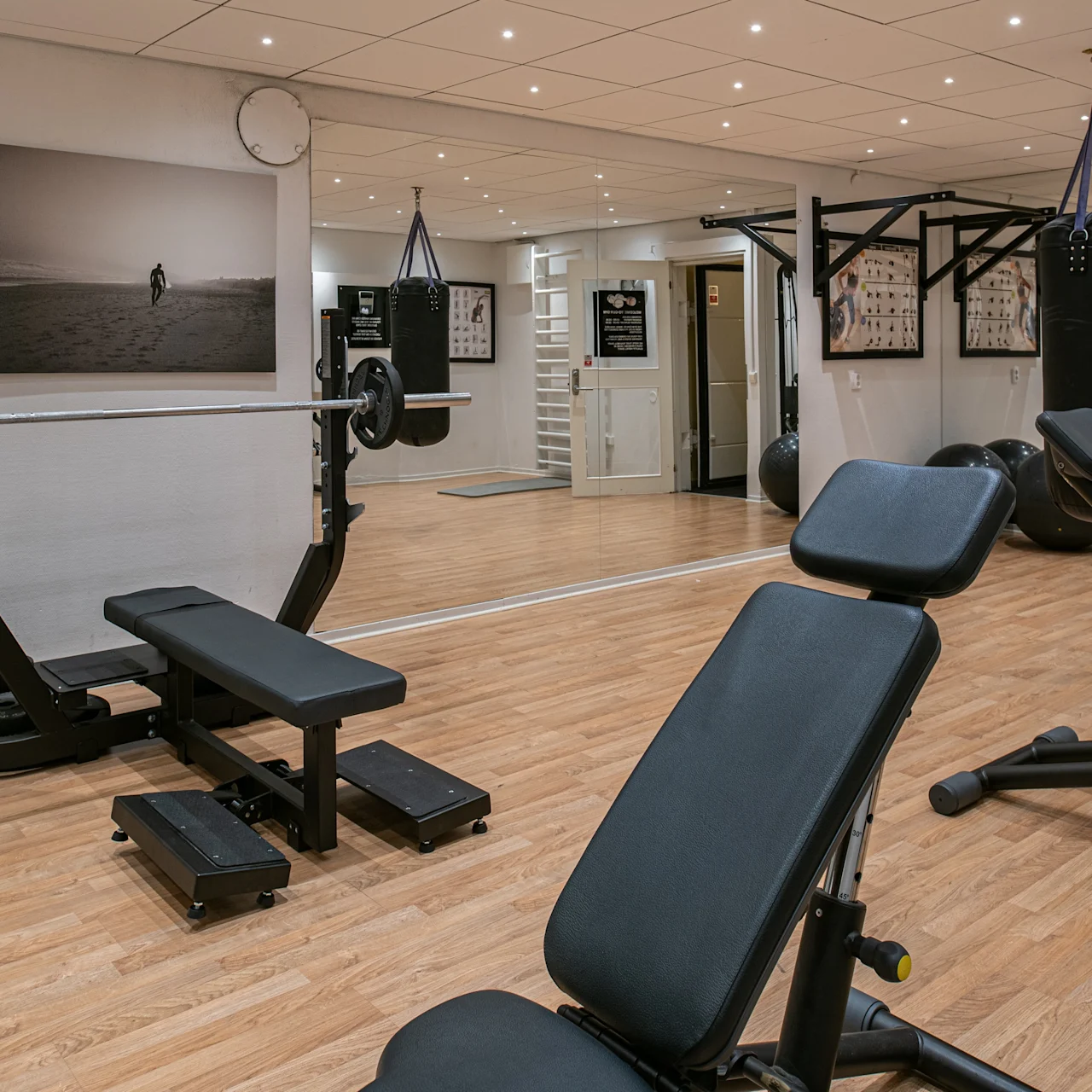 Vægtbænk i fitnesscenter omgivet af træningsudstyr på trægulv. Spejle reflekterer pladsen, mens væggene viser træningsplakater og en boksebold hænger i nærheden.