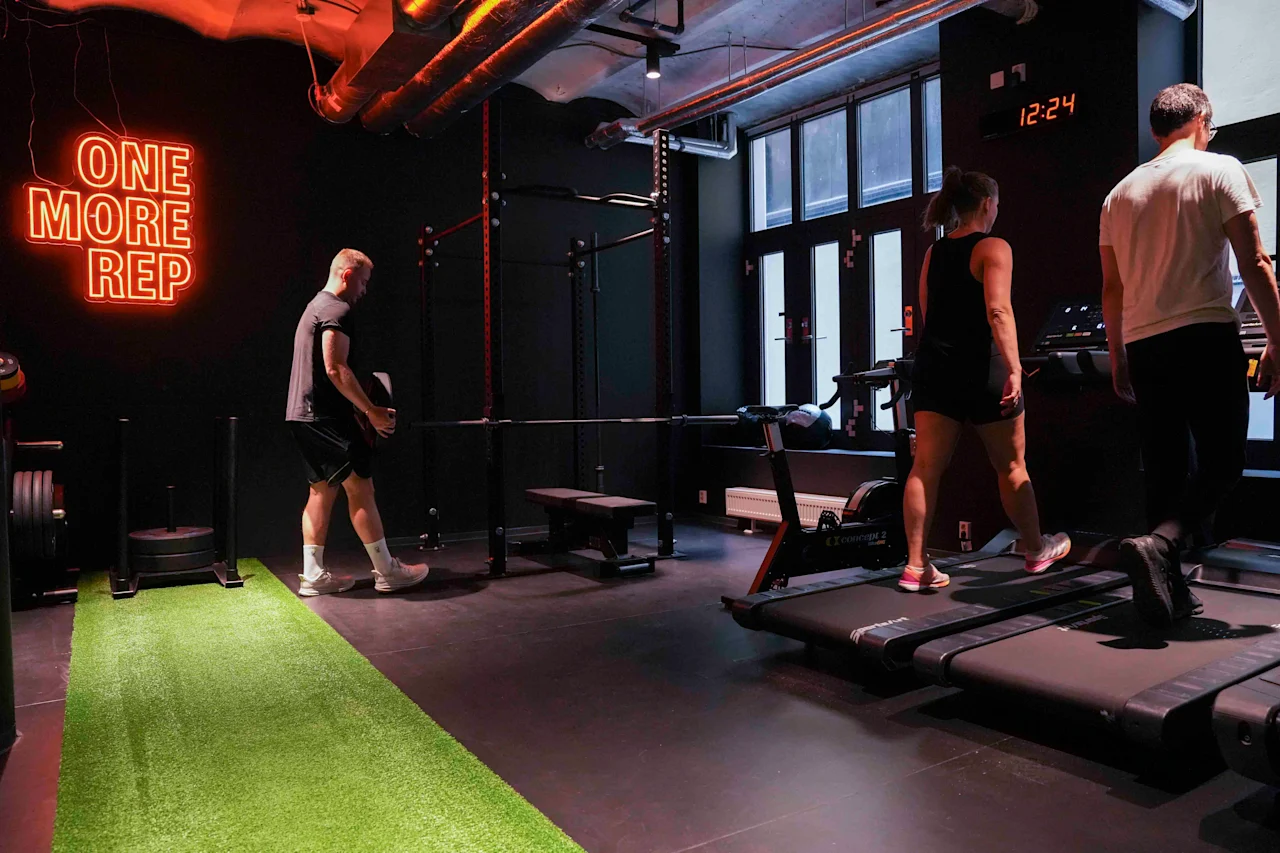 Et fitnesscenter med mennesker, der træner på løbebånd og en græsstribe, med et neonskilt, der siger "ONE MORE REP" og et ur, der viser 12:24.