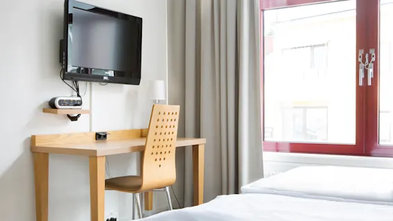 En veggmontert TV ser mot et skrivebord av tre og stol i et minimalistisk rom. Store vinduer med rødt ramme og hvite gardiner slipper inn naturlig lys.