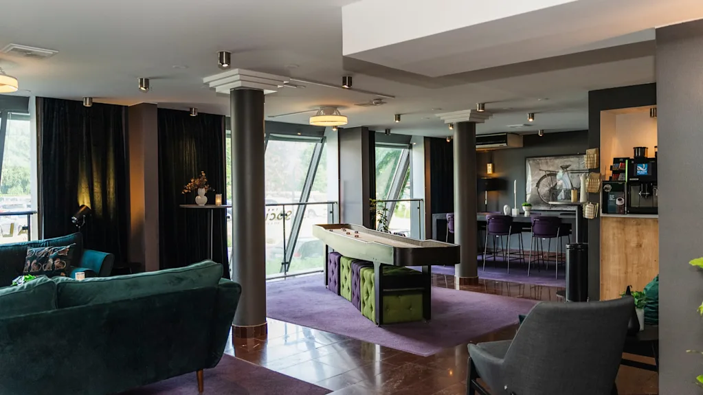 Ett modernt sällskapsrum med ett shuffleboardbord omgivet av bekväma sittplatser och samtida inredning. Stora fönster ger naturligt ljus, med en mysig sittgrupp och en kaffemaskin i närheten.