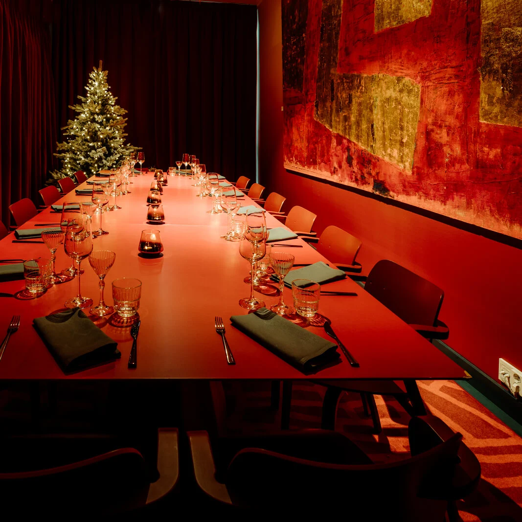 A long red dining table set with glassware and napkins, surrounded by chairs. Lit candles adorn the center, with a Christmas tree in the background. Dark red curtains provide ambiance.