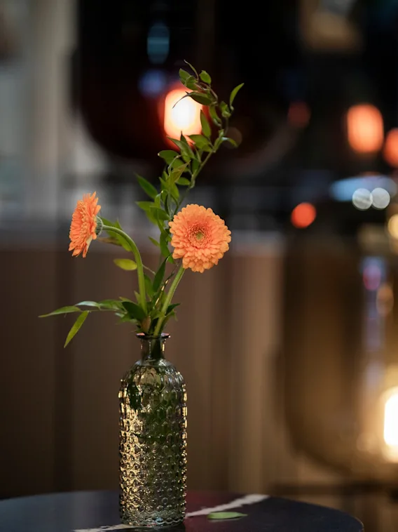 En koselig kveldsscene på Home Hotel Grand Bodø med en vase med oransje blomster på et bord og varme, uskarpe lys i bakgrunnen.