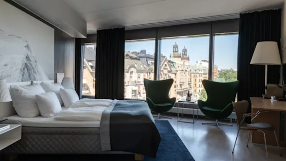 A modern hotel room at Clarion Hotel Sign with a comfortable bed, two armchairs, and a city view from a large window.