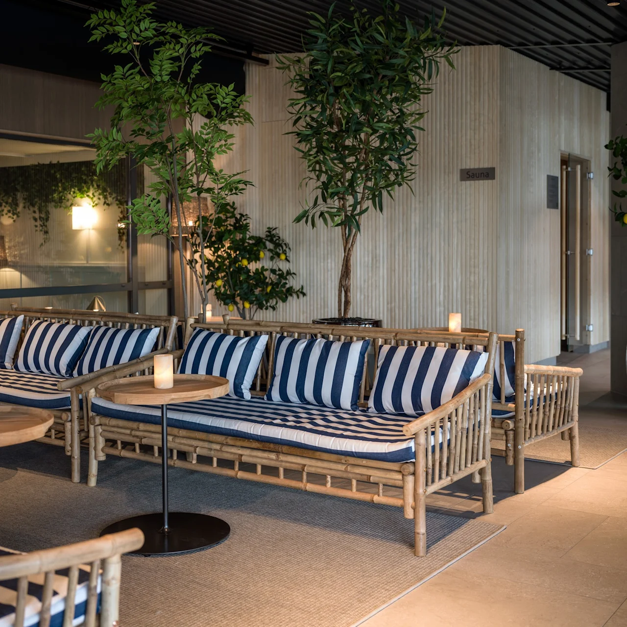Träsoffor med blå- och vitrandiga dynor står i en lounge, omgivna av krukväxter. En skylt på väggen säger "Sauna." Ljus på borden skapar en varm atmosfär.