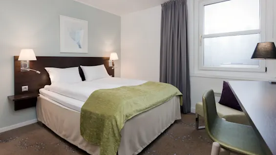 A neatly made double bed with a green throw rests under wall lamps, in a modern, softly-lit room with a large window and minimalistic decor.