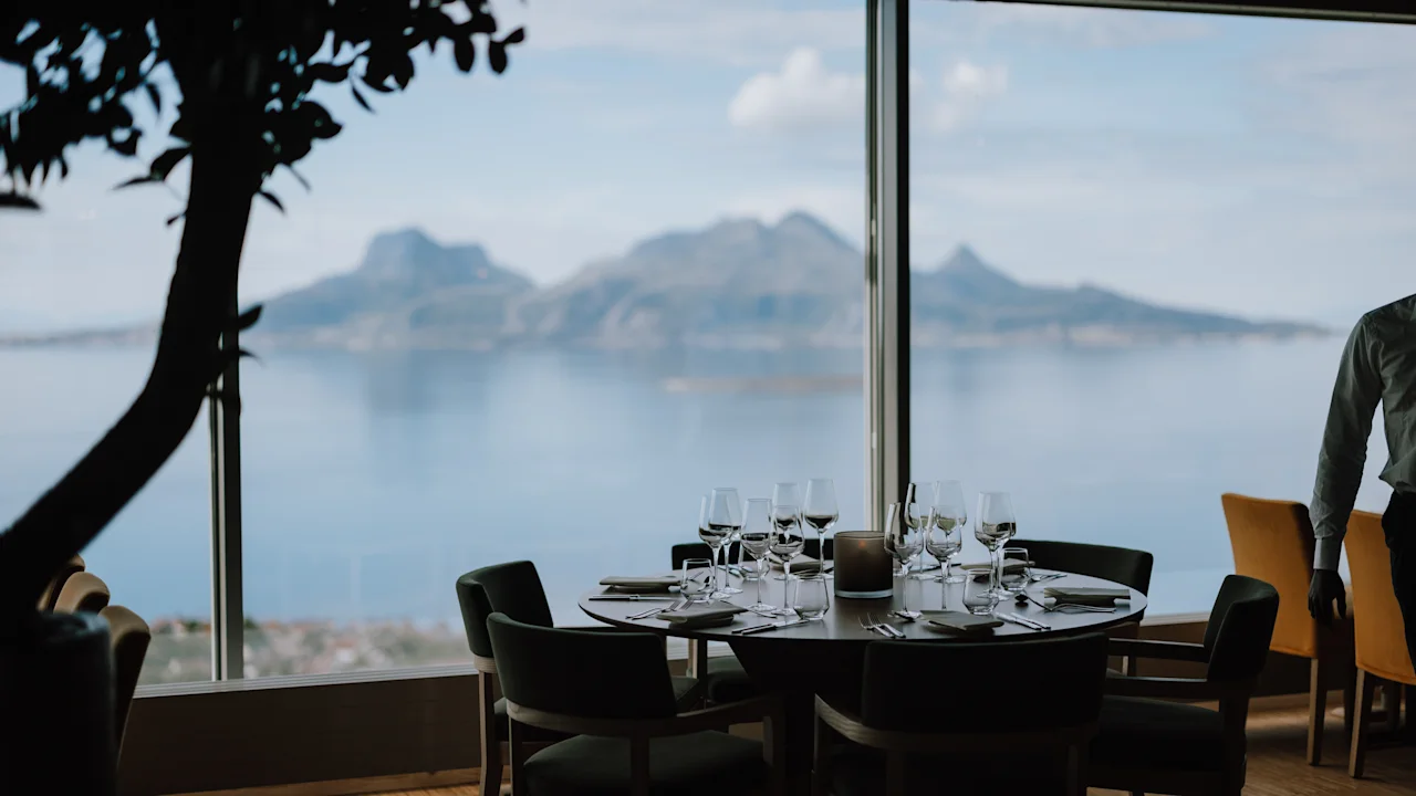 Et bord med pent ordnede glass og bestikk har panoramautsikt over fjell og vann gjennom et stort vindu. En person står i nærheten i en restaurantsetting.