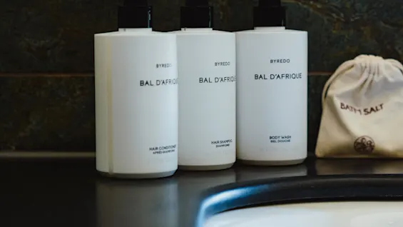 Tre hvite kosmetikkflasker merket "BYREDO BAL D'AFRIQUE" for balsam, sjampo og kroppsvask, ved siden av en badesaltpose, på en mørk benkeplate ved siden av en hvit vask.