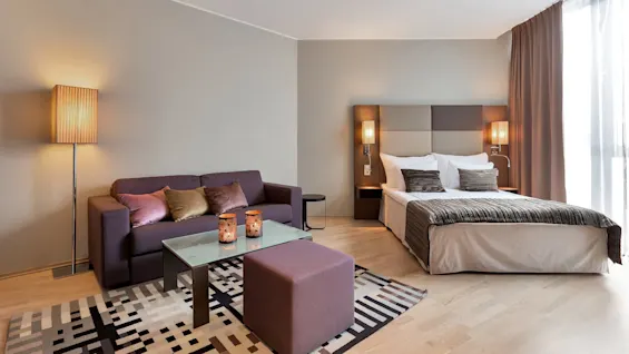 A hotel room with a large bed, patterned cushions, and a glass coffee table, featuring modern lighting and warm, neutral colors, creates a cozy and welcoming atmosphere.