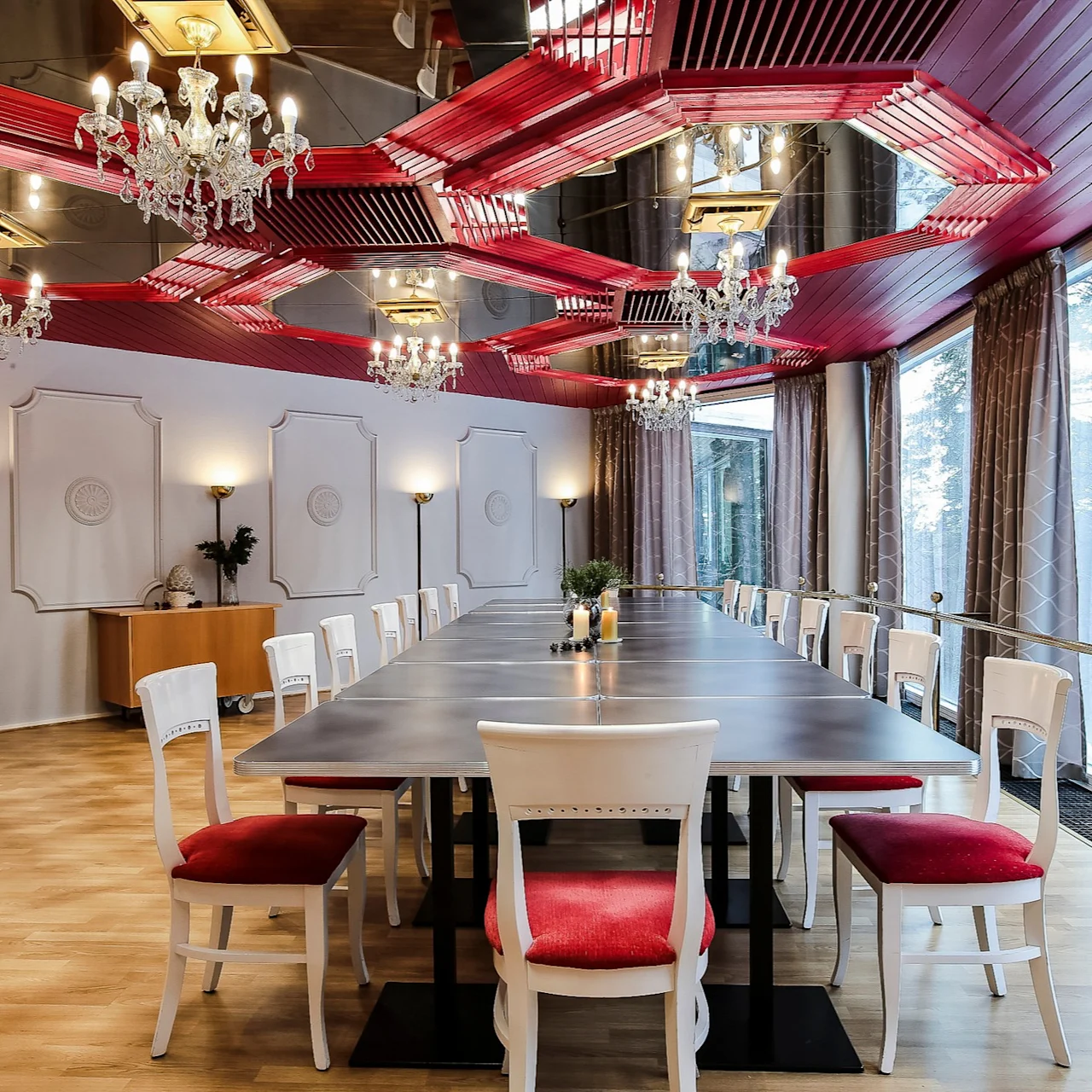 A long dining table with white chairs featuring red cushions is set in an elegant room. Chandeliers hang from a red and mirrored ceiling; large windows on the right offer a wooded view.