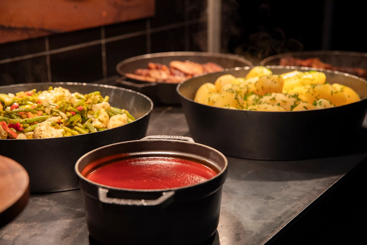 En varm buffé med ulike retter i svarte gryter, inkludert dampede grønnsaker, poteter med urter og en fyldig rød saus.