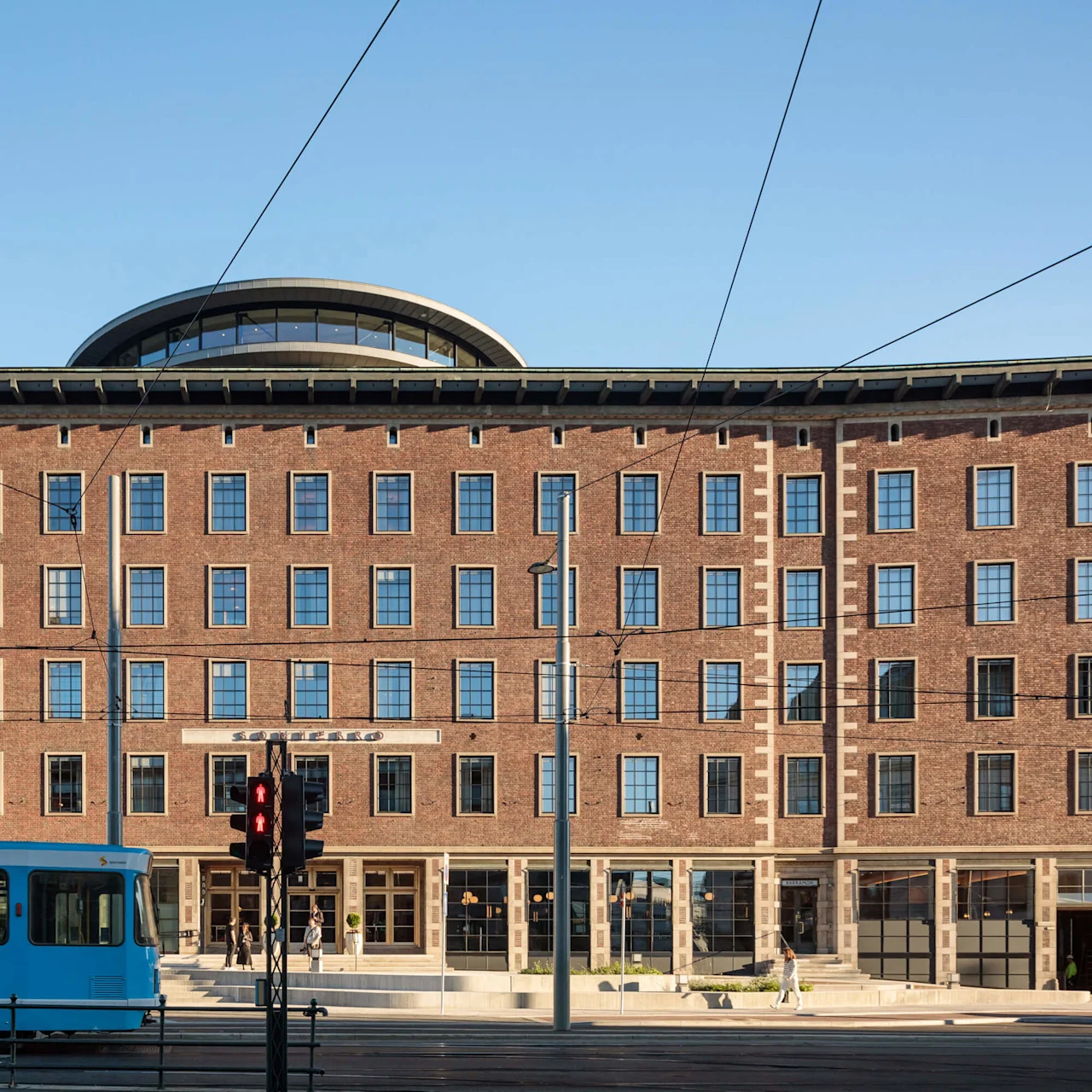 En murstensbygning med store vinduer bag en sporvogn på skinner; klar himmel.