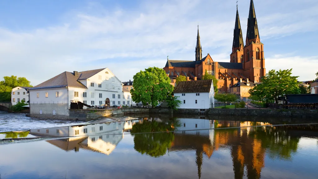 A scenic view of Uppsala with a river reflecting historic buildings and a grand cathedral under a blue sky.