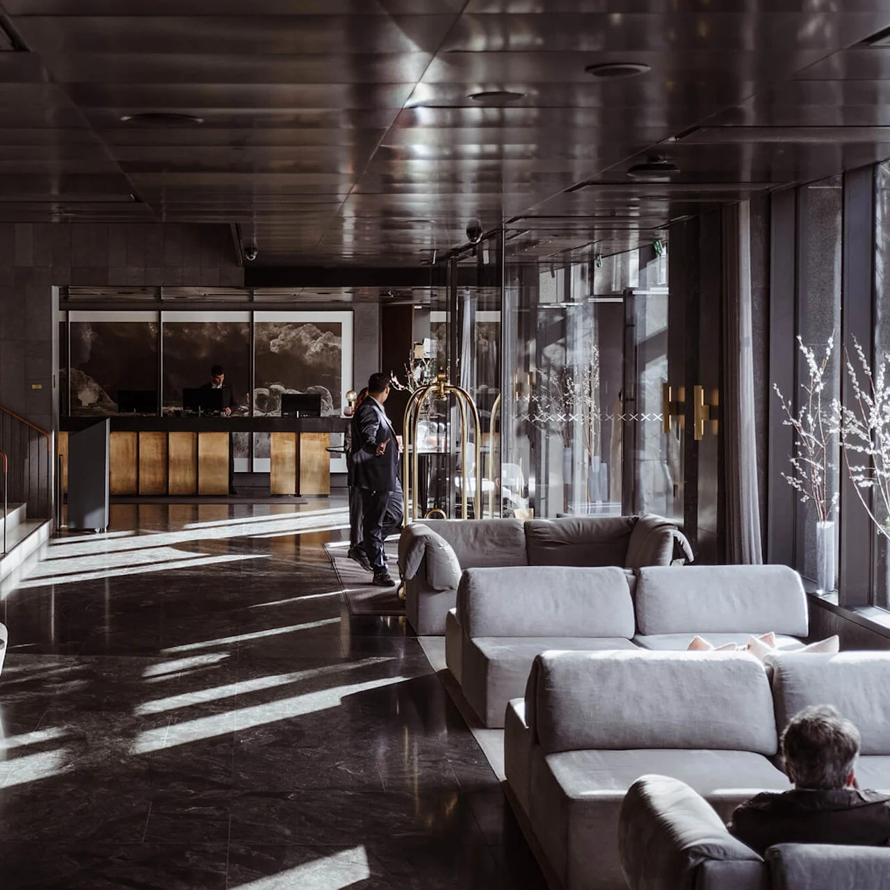 En hotellobby med en resepsjonsskranke; en person sjekker inn. Myke sofaer og lenestoler fyller det solbelyste rommet, med moderne dekor og utsikt fra vinduer.