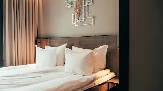 A cozy hotel room with a neatly made bed, white pillows, a wooden headboard, and a modern wall decoration.