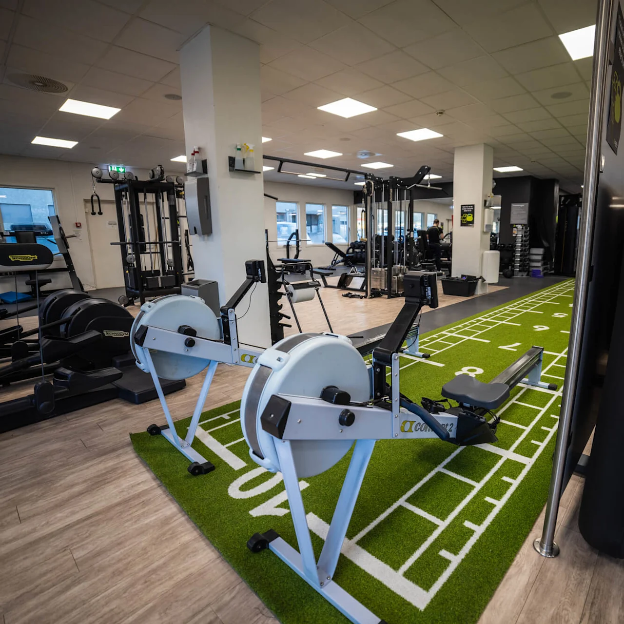 Romaskiner placeret på et grønt kunstgræs på banen inde i et rummeligt træningscenter med forskelligt fitnesudstyr. Området er godt oplyst med lysstofrør og store vinduer.