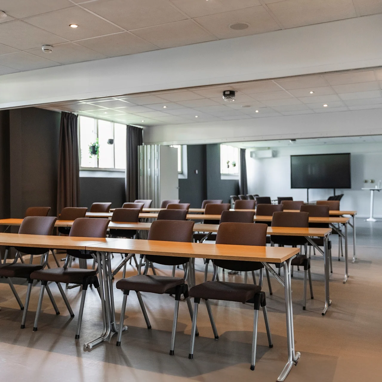 Rader av tomma bord och bruna stolar är prydligt arrangerade i ett ljust, modernt konferensrum; en stor skärm är synlig längst fram.