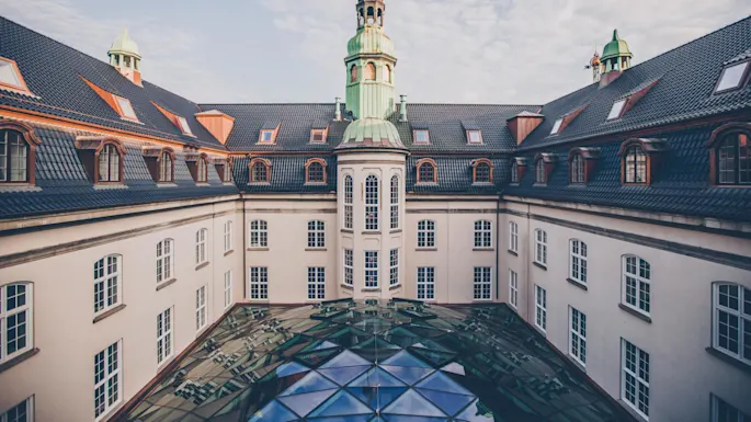 An ornate building with a central tower and glass dome roof. The building features multiple windows and blue tile roofing. The sky is partly cloudy.