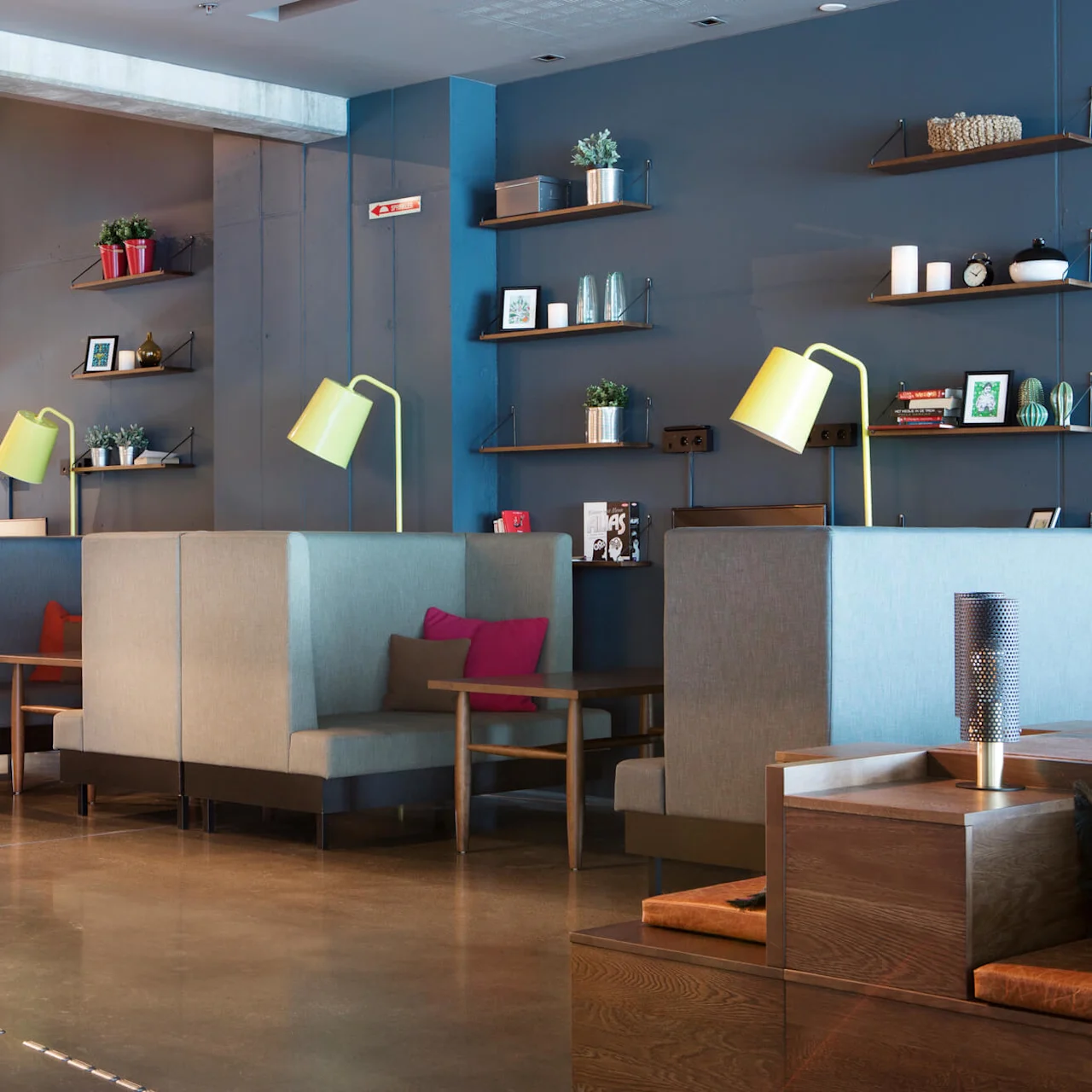 Grå båser med trebord og gule lamper skaper en moderne loungeatmosfære. Hyller på en blå vegg har planter, bilder og bøker. En rosa grafikk dekorerer sideveggen.