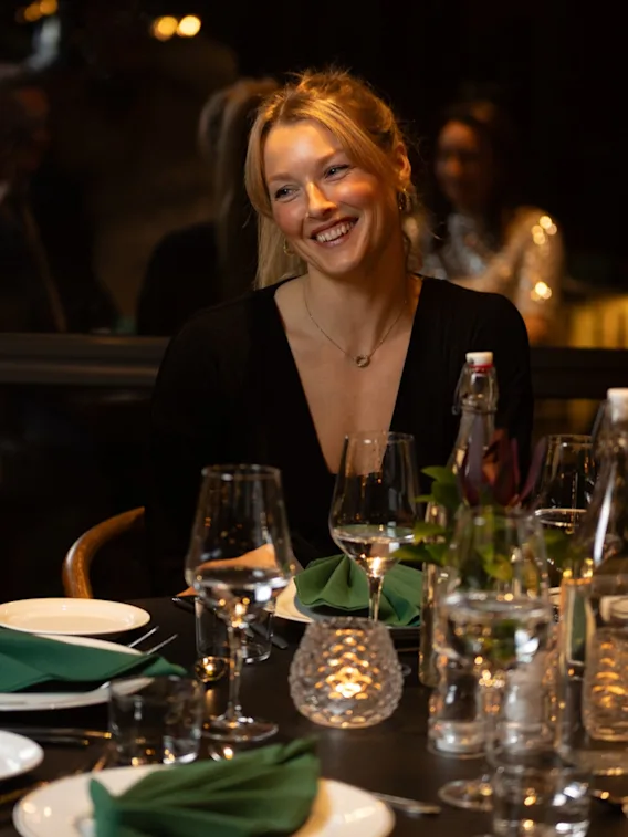 En person smiler lyst ved et middagsbord, og nyter et sosialt samvær på en restaurant.