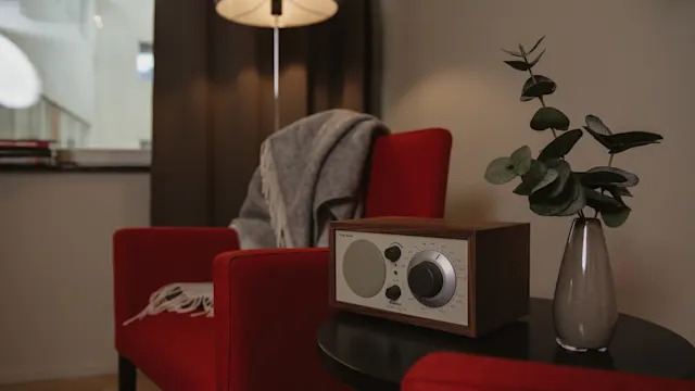 En vintage radio står på ett runt bord bredvid en vas med lövverk. Röda fåtöljer och en golvlampa skapar en mysig vardagsrumsatmosfär, med en filt hängande över en stol.