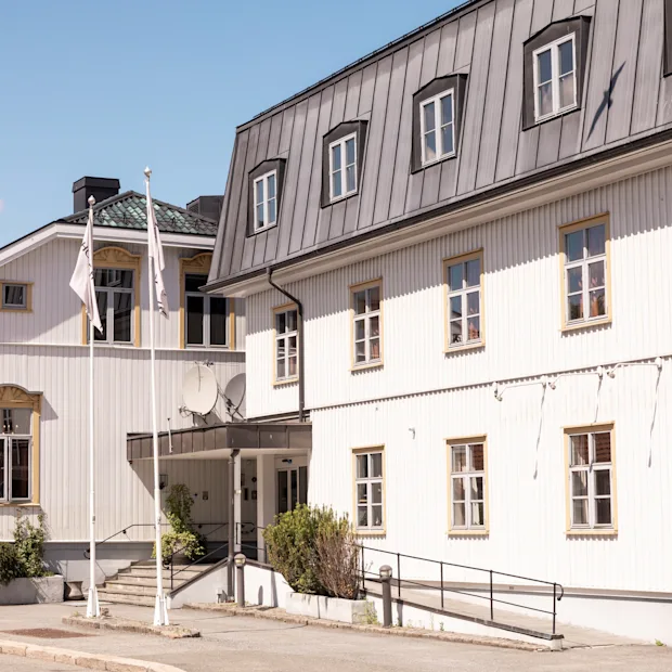 En hvid, toetagers træbygning med flere vinduer og metaltag befinder sig på en solrig gade med klar blå himmel. Ingen tekst er synlig.
