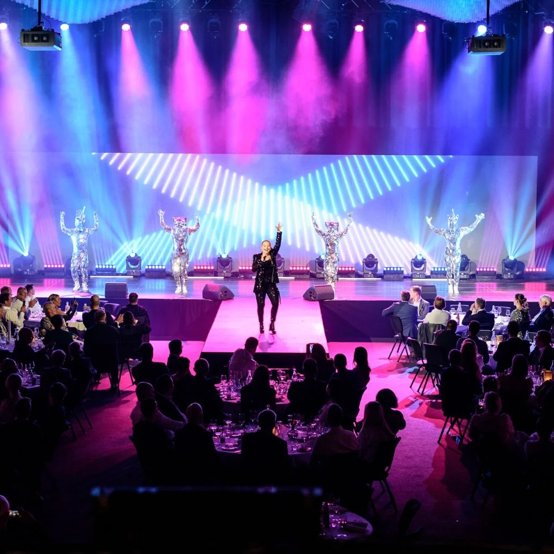 A performer sings on stage with colorful lights and dancers, while an audience sits at tables in a dimly lit event space.