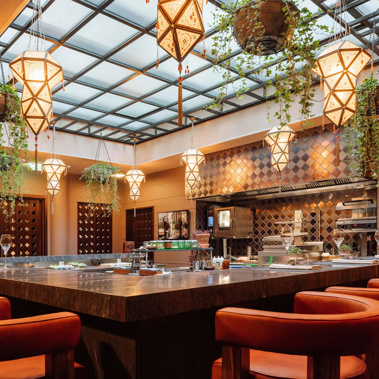En elegant bar med læderstole, geometriske hængende lamper og planter, placeret under et ovenlysvindue i et varmttonet rum.