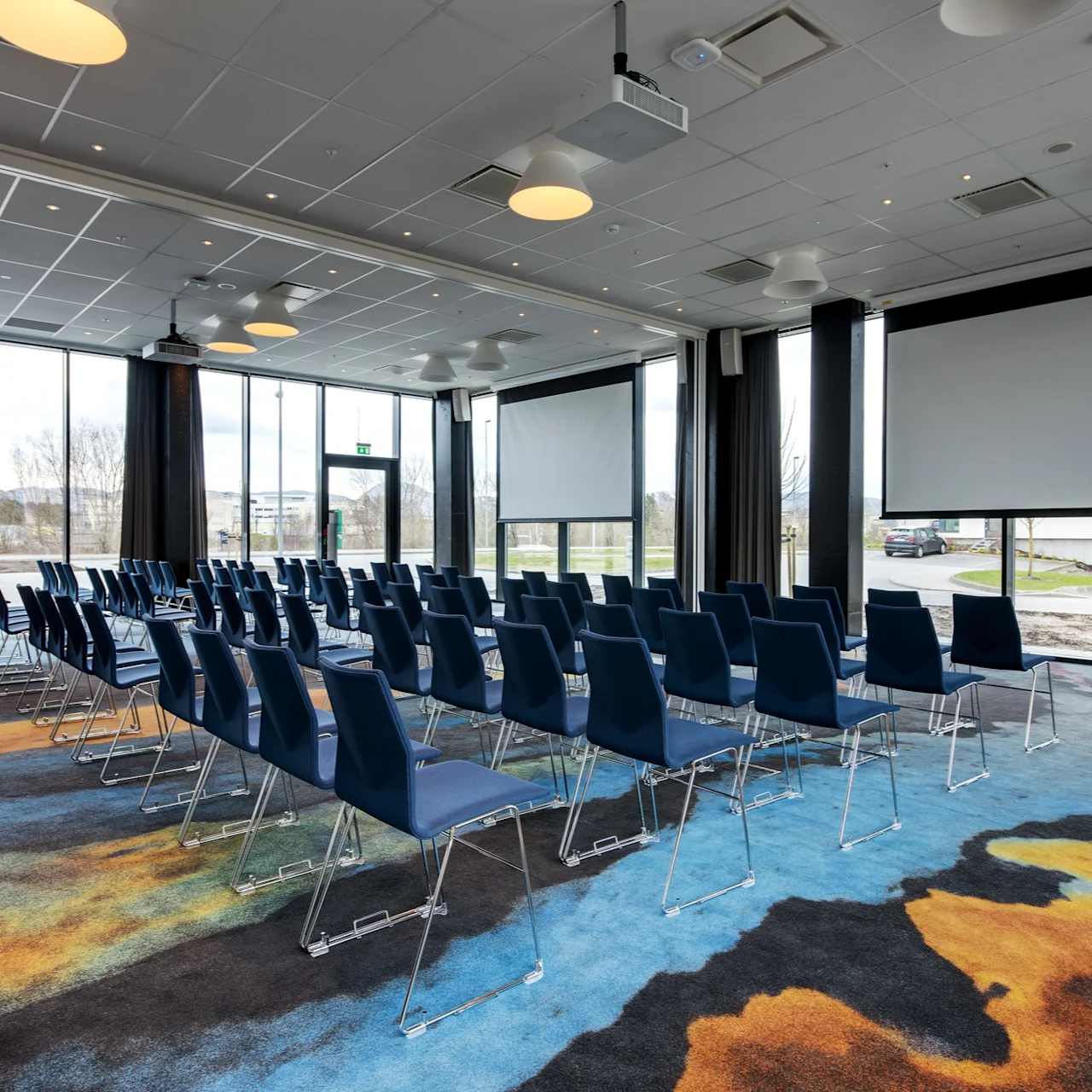 A conference room features rows of empty blue chairs facing two large projection screens, with large windows providing natural light and a colorful carpet beneath.