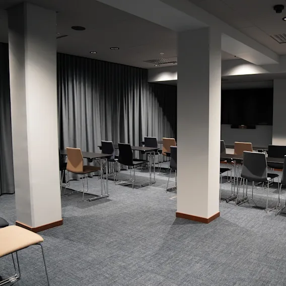 Rader av tomma stolar och bord är arrangerade i ett konferensrum. Rummet är svagt upplyst, med gråa gardiner längs väggarna.