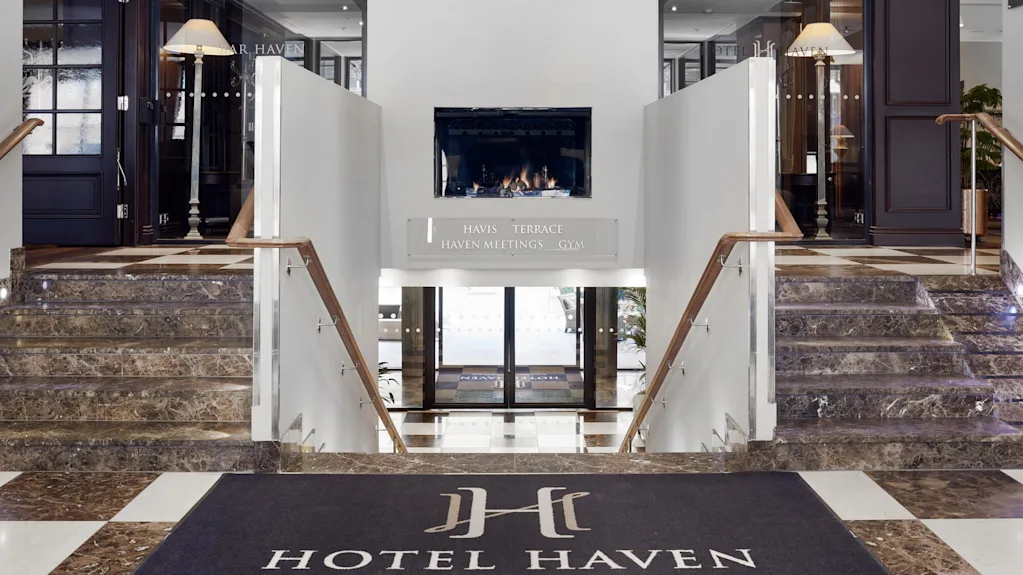 Entréhall med marmortrapper, der fører ned til glasdøre. En pejs er centralt placeret ovenover, flankeret af dekorative lamper. Skilt siger "Havis Terrace Haven Meetings Gym" og måtten siger "Hotel Haven."