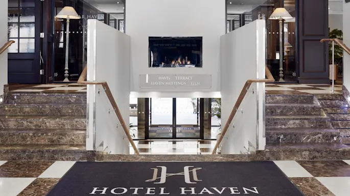 Entréhall med marmortrapper som fører ned til glassdører. En peis er sentrert over, flankert av dekorative lamper. Skiltet sier "Havis Terrace Haven Meetings Gym" og matten sier "Hotel Haven."
