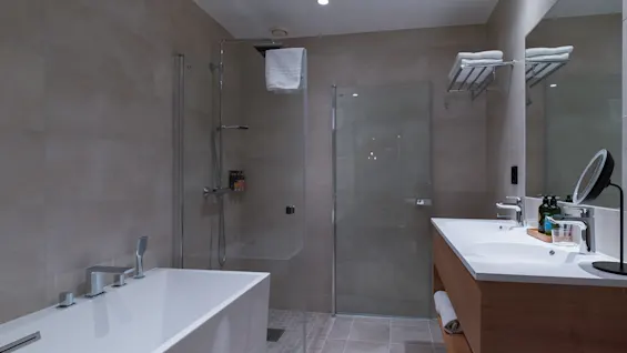 Freestanding bathtub sits unused beside glass-enclosed shower; sink with mirror and toiletries on wooden cabinet in modern bathroom with beige tiles.