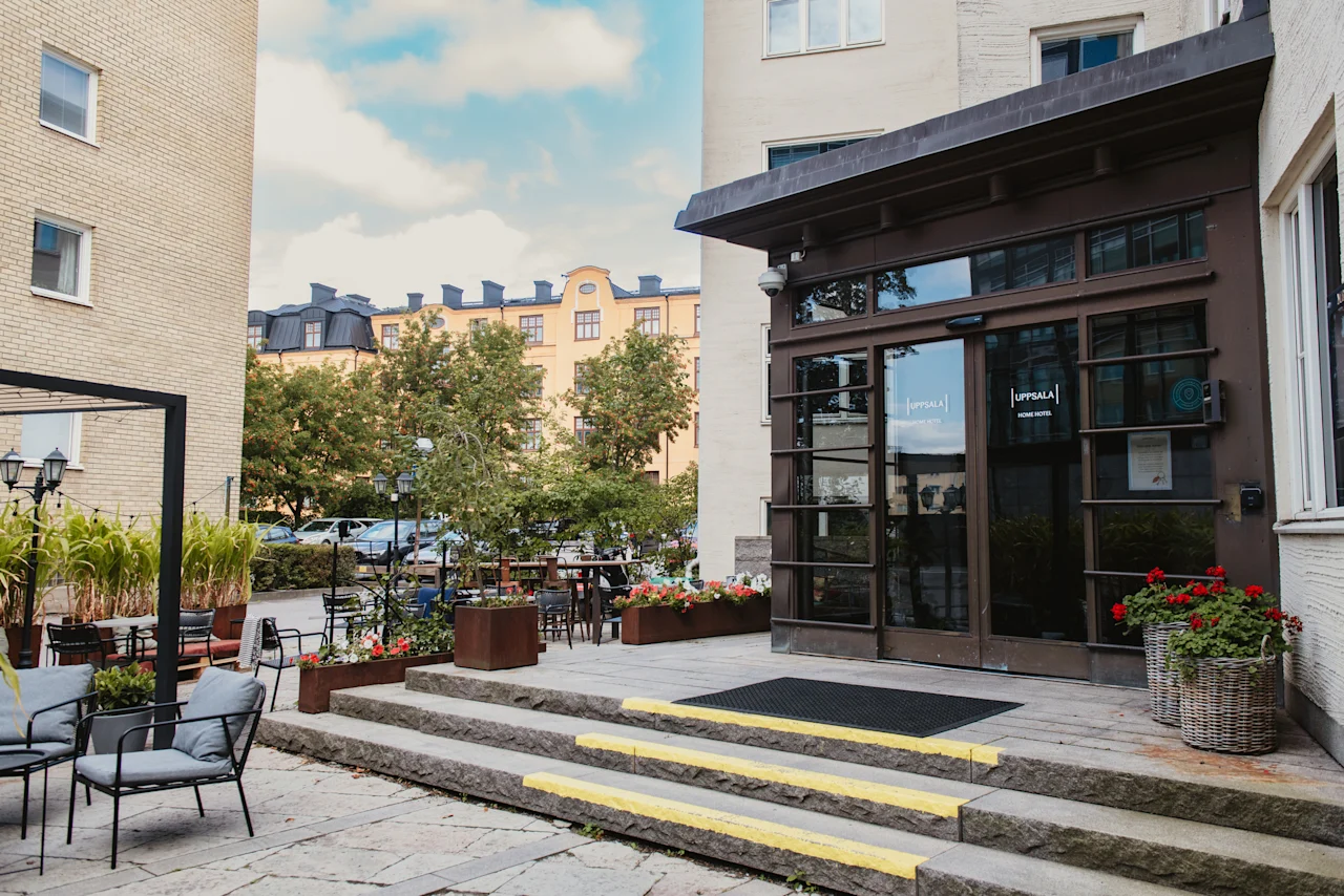 Outdoor seating area and entrance to Uppsala Home Hotel on a sunny day with trees and buildings in the background.