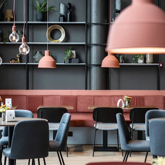 Runde bord med blomster er arrangert i en kafé med moderne stoler, rosa hengelamper og en hylle dekorert med planter og skulpturer.