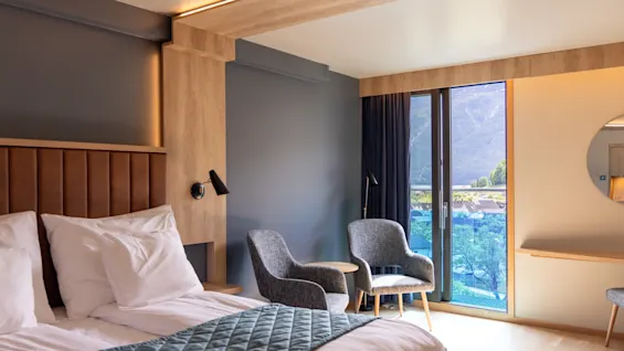 En säng med vita lakan och ett vadderat överkast ligger bredvid grå stolar. Stora fönster visar en utsikt över bergen och skapar en mysig hotellrumskänsla.