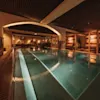 A rectangular indoor pool with gentle lighting, surrounded by lounge seating and wooden beams, offers a warm and inviting atmosphere for relaxation.