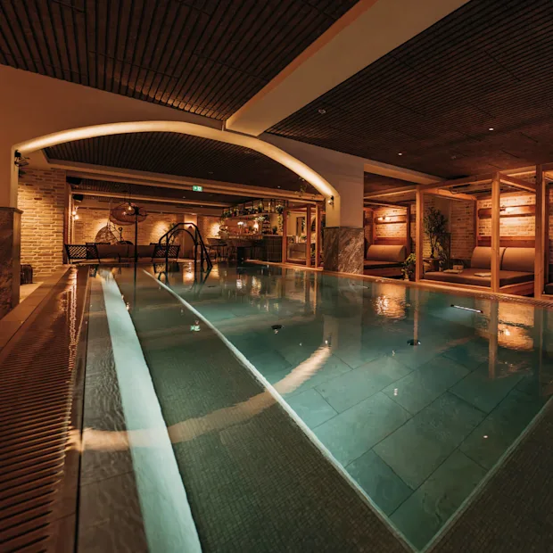 A rectangular indoor pool with gentle lighting, surrounded by lounge seating and wooden beams, offers a warm and inviting atmosphere for relaxation.