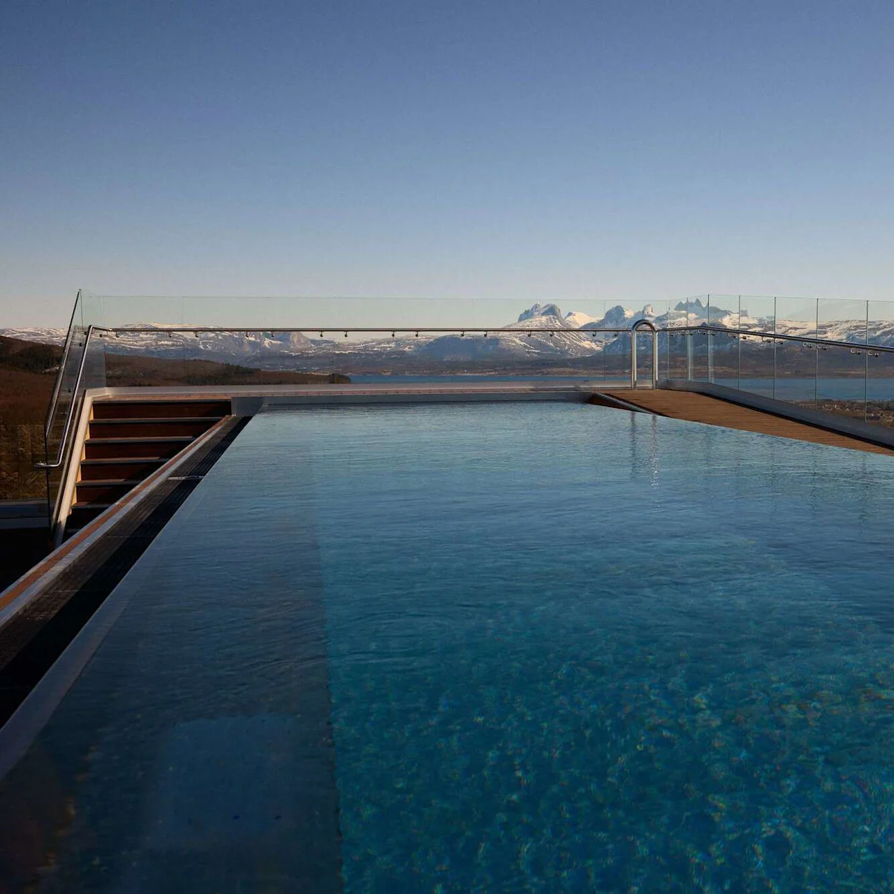 Infinity pool extends towards glass railing, overlooking snow-capped mountains under a clear blue sky.