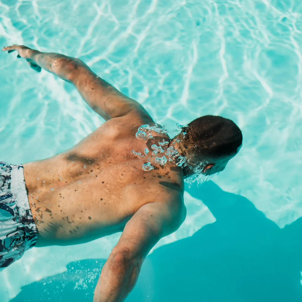 A person swimming underwater in a bright blue pool, with sunlight dappling the water's surface.