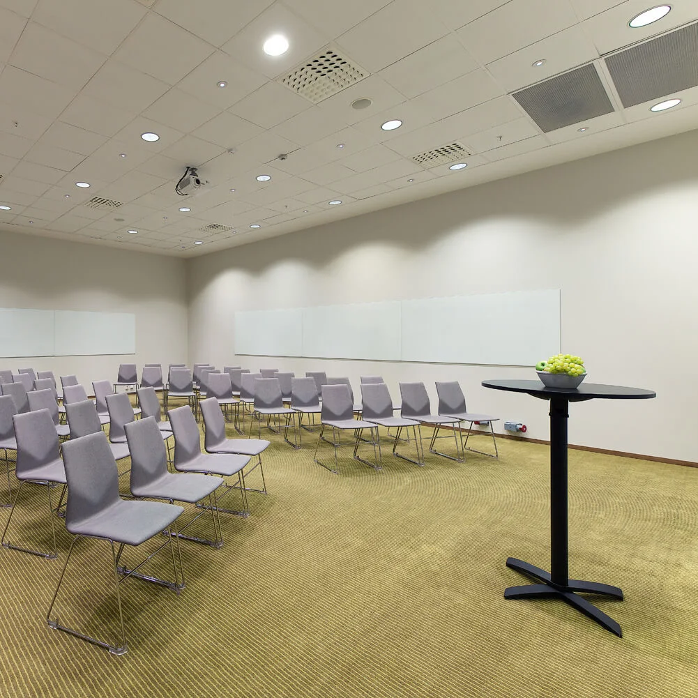 Rows of empty grey chairs face a blank whiteboard in a carpeted conference room; a small table with green grapes stands nearby.